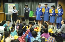 エプロン姿の6人の関係者が正面に立って話をしており、向かいに座っている多くの子ども達が手を挙げている写真