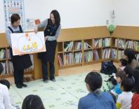 2人の関係者がひよこのイラストが入った紙を見ながら読み聞かせをしており、多くの親子連れの来館者が座って見ている写真