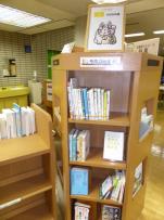 図書館の一角に、上部に猫のイラストが置かれた柱状に独立した本棚が設置されており、4段になった本棚に様々な図書が展示されている写真