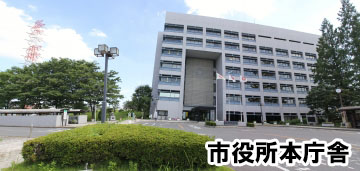 グレーを基調とした7階建てのビルの前に、三郷市の市章の旗が3つ掲げられ、駐車場になっている正面玄関の写真