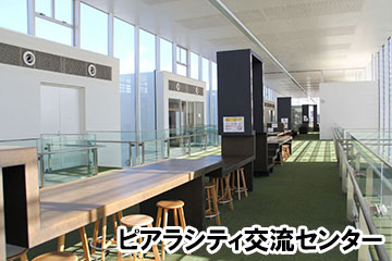 廊下の中央に木製の机と椅子が並べられ、一定感覚に黒い門のような入口があるピアラシティ交流センター内の写真