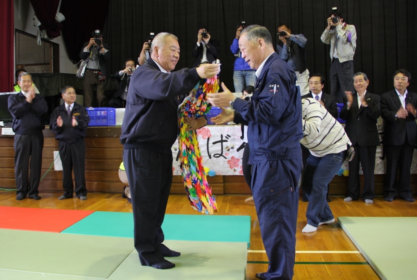 体育館の舞台に沢山のカメラマンがおり、市長から千羽鶴を受け取る山田町長の写真