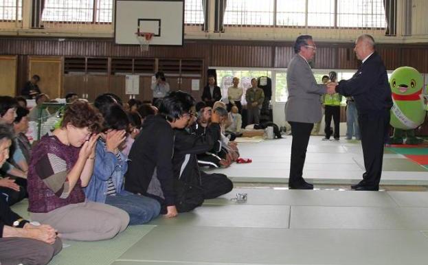 体育館の中で避難者の方々が座り、前で握手する木津市長と町議会議員の写真