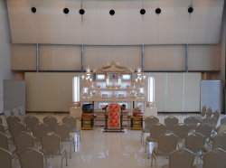 ベージュ系の色で統一された室内に、通路を空けて両側に椅子が数列並べられており、通路の先中央に電気の灯った白木の祭壇が置かれている本館の写真
