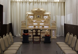 白い壁にダークブラウン立ち上がりと床の室内に、両壁に沿って椅子が対面に並べられ、その奥に白いカーテンを背に白木の祭壇が置かれている新館の写真