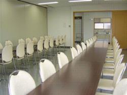 白い壁と床の室内に、奥に長く2列並んだ長机に、それぞれ白い椅子が複数対面に並べられている待合室の写真