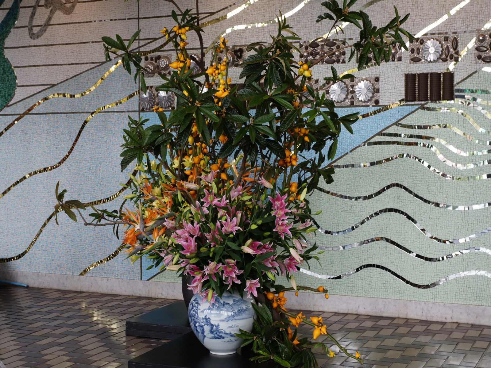 金色の波線模様が描かれた壁の前に、円形の花瓶にピンク色をしたユリやオレンジ色の花などが飾られている写真