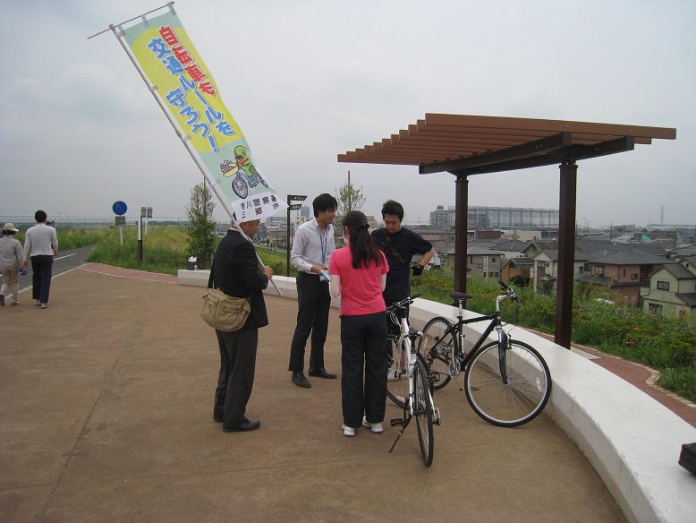 江戸川サイクリングロード（みさとの風ひろば）でスーツを着た男性が「自転車も交通ルールを守ろう！」というスローガンの下にかいちゃんが自転車に乗ったイラストが描かれた旗を持ち、もう一人の男性がサイクリングに来ていた男性と女性に自転車の安全利用を呼びかけている写真