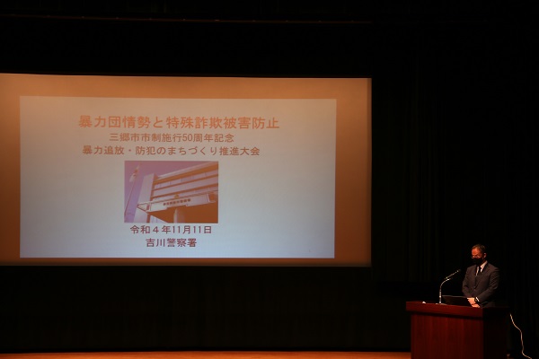 スクリーンに「暴力団情勢と特殊詐欺被害防止」の内容が映し出され、右側の演台前でマイクに向かって話をしている講師の男性の写真