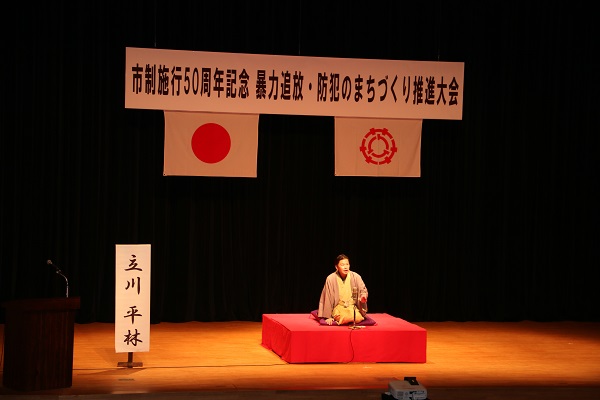 ステージ左側に、「立川 平林」と書かれためくりが置かれ、ステージ中央に設置された高座に座り落語を演じている立川 平林氏を客席から写した写真
