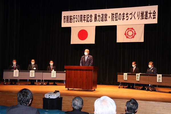大会の横断幕と、国旗・市章が掲げられたステージで左右に置かれた長机に副市長や講師らの関係者が着席し、中央の壇上であいさつをする市長の写真