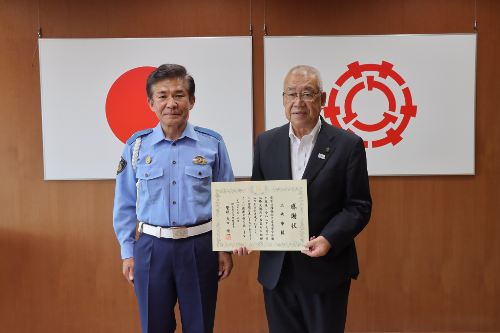 壁に掛けられた日本国旗と三郷市市章の前に立つ警察官の男性と感謝状を持つ三郷市長の写真