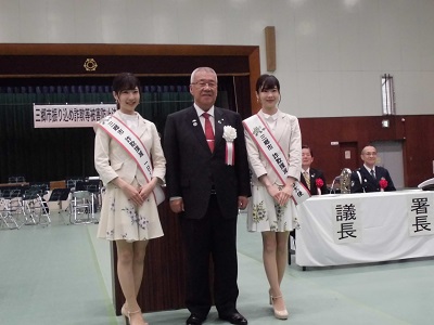 スーツを着て胸章を付けた市長とその両脇に立つ一日大使のたすきを付けた、Mika+Rikaのお二人の写真