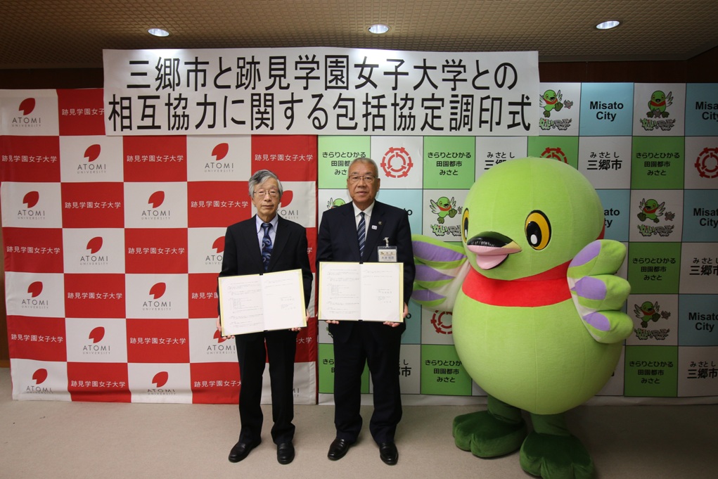 山田跡見学園女子大学学長と木津三郷市長が協定書を持っており、市長の右側に三郷市キャラクター「かいちゃん」が立ち、一緒に記念撮影をしている写真