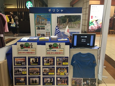 テナントが並ぶ施設内の一角に「ギリシャ」と書かれたイベントコーナーが作られ、青いTシャツやホストタウン交流の写真がボードに展示されている写真