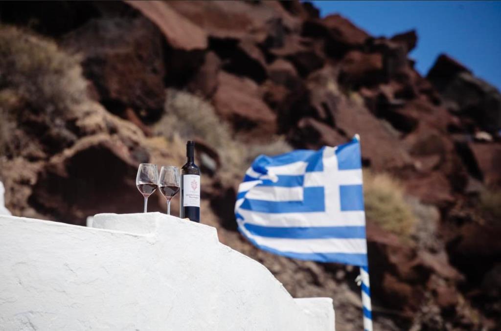 風でなびいているギリシャ国旗の左手前にワインが注がれた2個のグラスと瓶が並んでいる写真