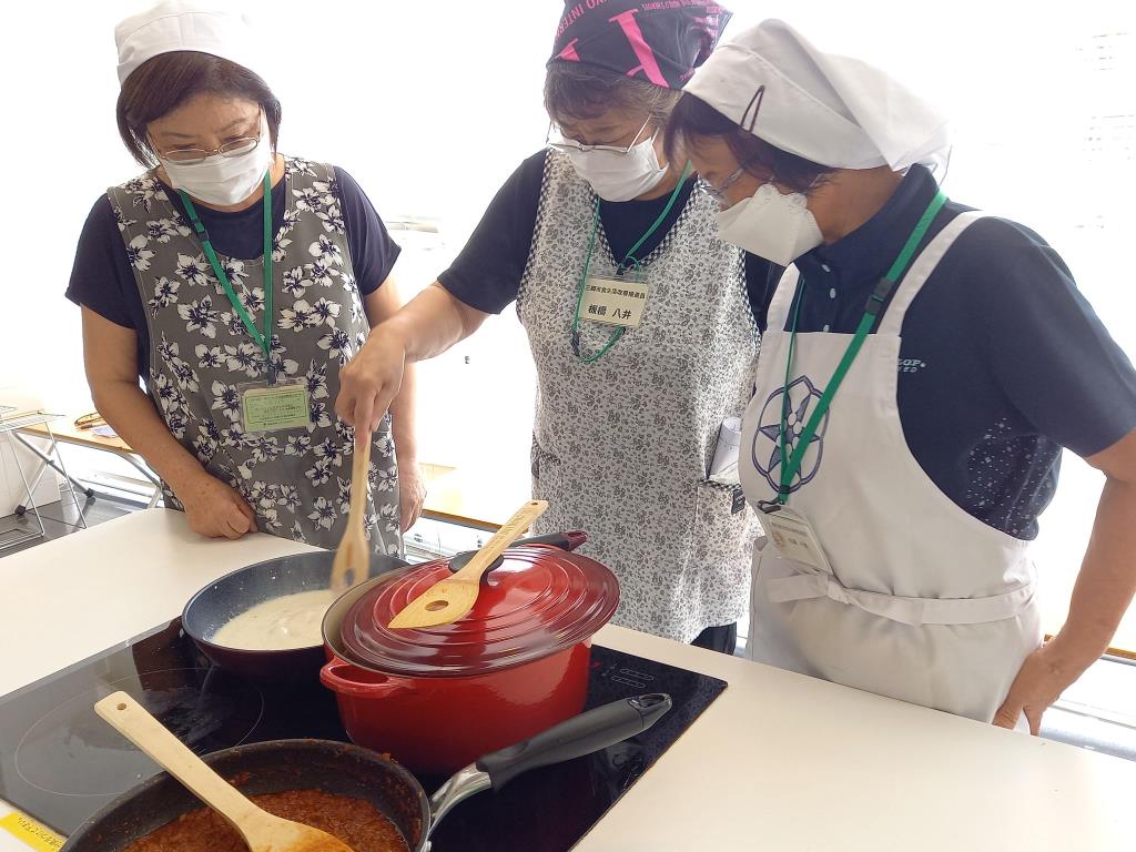 三角巾にエプロンを着用した女性が煮詰めた鍋を混ぜている様子を2名の女性が見ている写真