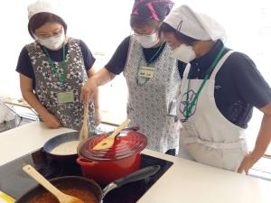 三角巾にエプロンを着用した女性が煮詰めた鍋を混ぜている様子を2名の女性がそばで見ている写真