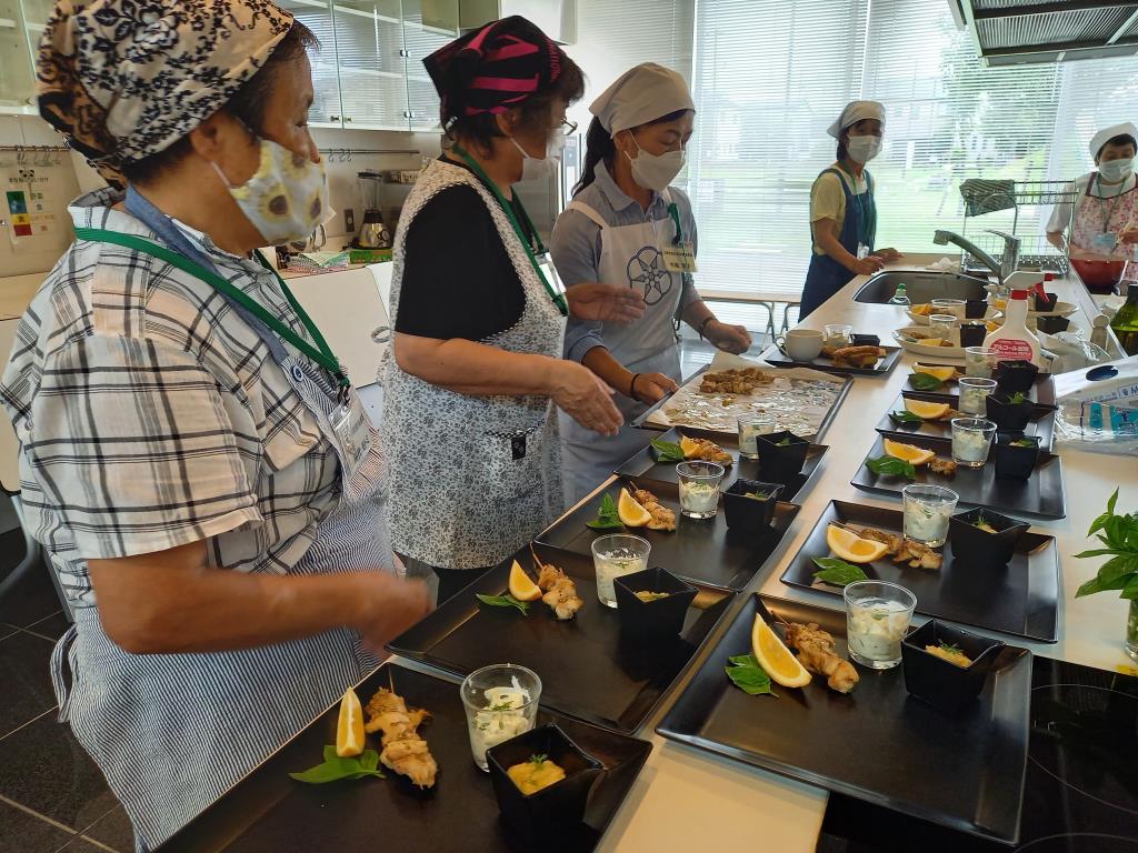 三角巾にエプロンを着用した女性がオーブンで焼き上げたムサカを切り分け完成した順番に黒色の四角形のお皿に盛り付けている様子を2名の女性が見ている写真