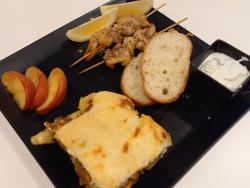 カットしたフランスパン2切れに肉の串焼き「スブラキ」が2本、カットしたリンゴ3切れとレモン2切れ、とろけるチーズに焼き目が付いて焼き上がった「ムサカ」、白いソース「ザジギ」が添えられ完成したギリシャ料理の写真