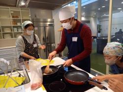 白色の三角巾に紺色のエプロンを着用した男性がフライパンで材料を炒めている写真