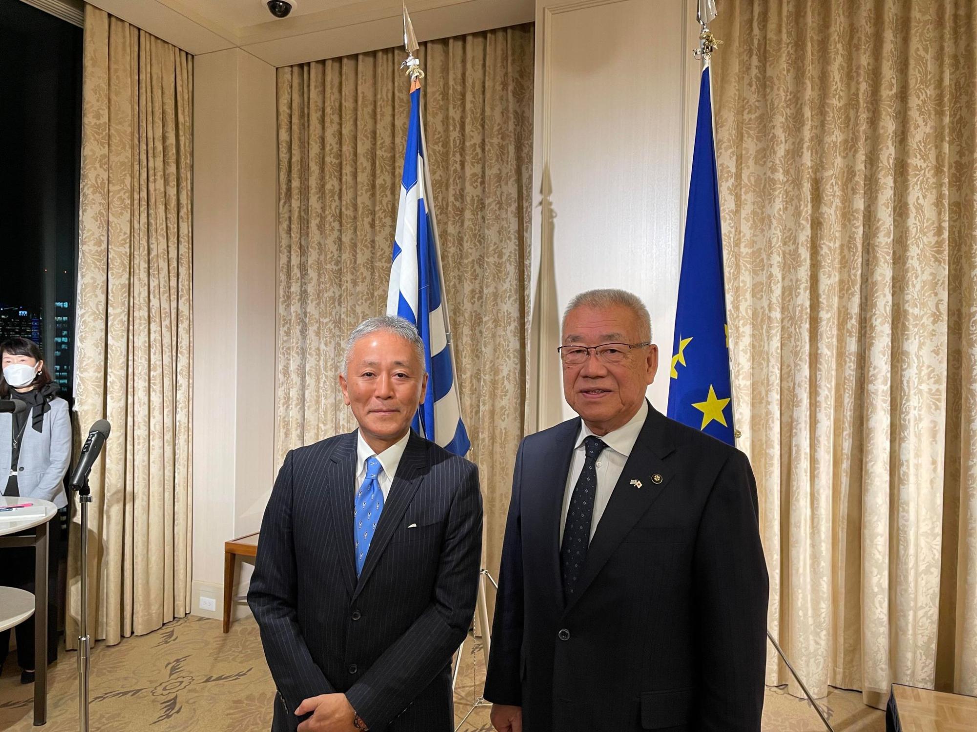 2本の国旗が立つ前で中山駐ギリシャ日本国大使と市長が並んで立っている写真