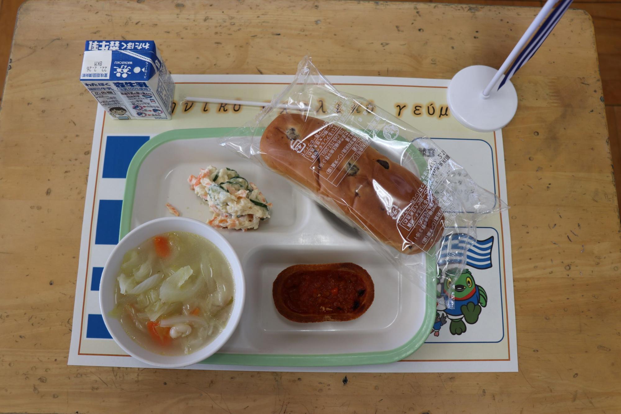 給食トレーに透明の袋に入ったぶどうパン、ムサカ、フムス入りサラダ、スープカップに入ったファソラーダが盛り付けられ、パックの牛乳が添えられた小学校のギリシャ給食の写真
