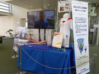 映像が流れているテレビの前に設置された青色のクロスが掛けられた台の上に聖火リレートーチやユニフォームなどが展示されている写真