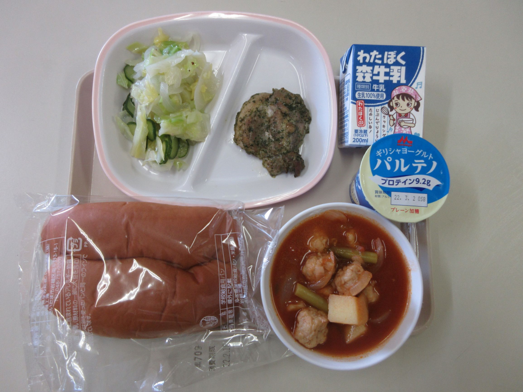 給食トレーに透明の袋に入った黒パン、スズカキア、ハーブチキン、グリークサラダが盛り付けられ、ギリシャヨーグルトとパックの牛乳が添えられた中学校のギリシャ給食の写真