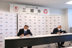 長机にJOCの山下泰裕会長と市長が間隔をあけて座り協定書にサインを行っている様子の写真