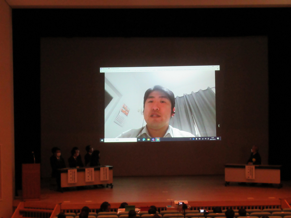 ステージ奥にリモートで挨拶を行っているギリシャ共和国在住の新井良様の顔が映されている写真