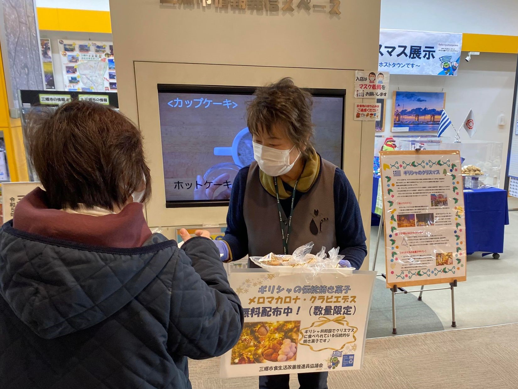 透明のビニール袋に入ったお菓子が入っている、ポップがついたトレイを持った女性スタッフがお菓子を貰いに来た女性客に配布している様子の写真