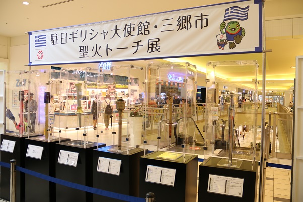 商業施設内の一角に「駐日ギリシャ大使館 三郷市 聖火トーチ展」のコーナーが作られ、様々な形をしたトーチがアクリルケースに展示されている写真
