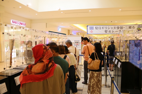 商業施設内の展示コーナーに様々な形をしたトーチがアクリルケースに入って展示されており、多くのお客さんで賑わっている写真