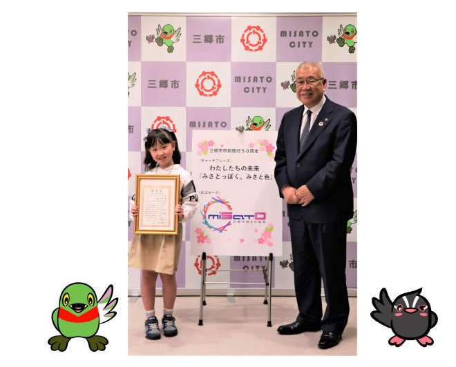 50周年キャッチフレーズの考案者 指宿彩音さん（左）が認定証を両手に持ち、市長（右）と一緒に三郷市のバックボードの前で記念撮影をしている写真とかいちゃんとつぶちゃんのイラスト