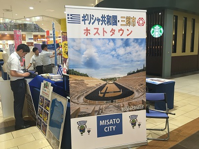 施設内の一角に「ギリシャ協和国 三郷市 ホストタウン」と書かれたイベントコーナーが作られ、関係者たちが展示物を準備している写真