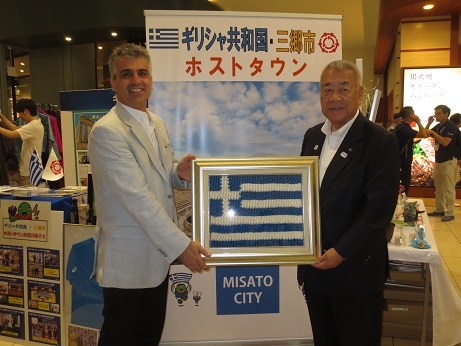 ホストタウンのイベントコーナーで駐日ギリシャ大使館 参事官と木津市長が一緒にギリシャ国旗の作品を持って記念撮影をしている写真