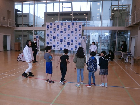 体育館で関係者が見守る中、子ども達が横一列に並んでボッチャの競技体験をしている写真