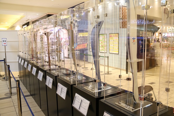 商業施設内の展示コーナーに様々な形をしたトーチがアクリルケースに入れられ展示されている写真