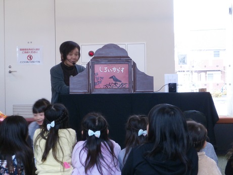 木枠に「しろいからす」と書かれた紙芝居が入っており、座って聞いている子ども達の前で紙芝居を上演する女性の写真