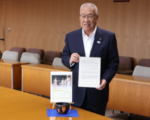 テーブルの上の東京2020オリンピック聖火採火式の写真の前に青色の敷き台が置かれ、敷き台の上に置かれた陶器の右側に手紙を両手で持っている市長の写真