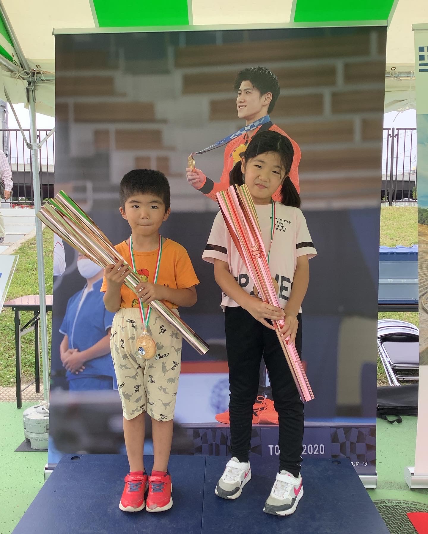 金メダルを首に掛けた選手の大きな写真パネルの前で女の子と男の子が横に並んで聖火リレートーチを両手で持っている写真