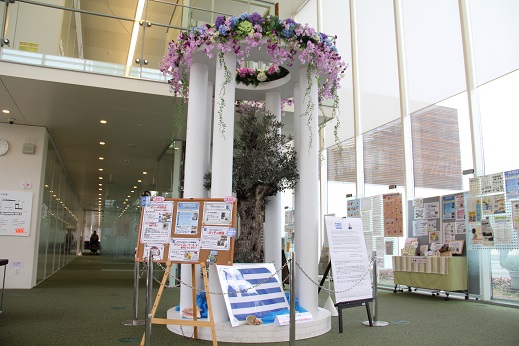 ガラス張りで開放的な空間に、木や花々が装飾されたモニュメントが設置され、横に置かれたボードとともに展示されている写真