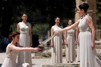 古代の衣装に身を包んだ3名の巫女の女性が見守るなか、左側の膝まづいた女性が持っている採火した火に右側に立っている女性がトーチを右手に持ち点火している採火式の様子の写真