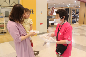 右側の女性スタッフが小さな女の子を抱っこした母親にギリシャの伝統菓子を配布している様子の写真