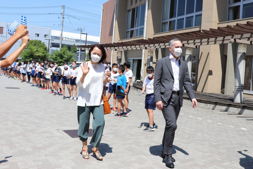 校庭に並んで立つ児童達に出迎えられ笑顔で歩いている駐日ギリシャ大使館職員2名の写真