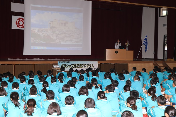 ギリシャ文化交流プログラムの横断幕が設置された体育館内で、プロジェクタースクリーンに映像を映しながら演壇で説明をしている関係者と、スクリーンを眺めている生徒たちの写真