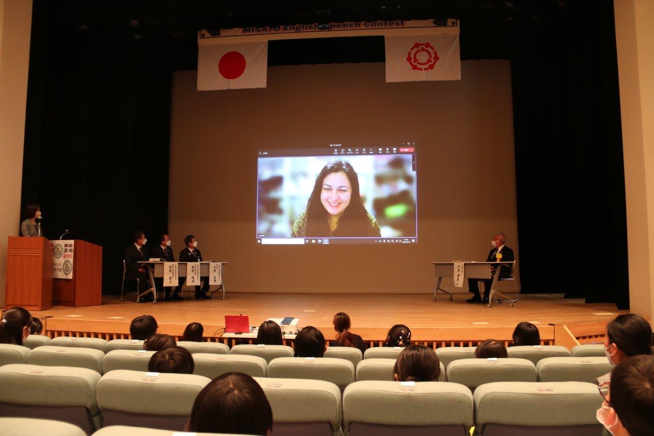 オンラインで審査に参加したアナスタシア・新井・カチャントニさんの挨拶の映像がステージ奥に映し出され、着席した参加者が聞いている写真