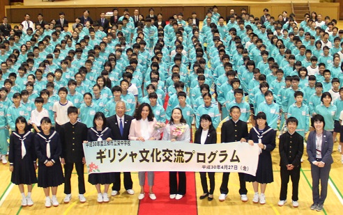 体育館でジャージ姿の栄中学校の生徒たちが整列している中、「ギリシャ文化交流プログラム」の横断幕を持って一緒に記念撮影をしている駐日ギリシャ大使夫人や木津市長の写真
