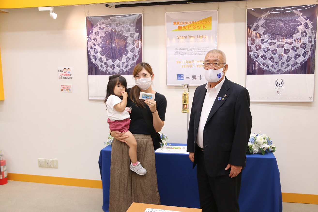 聖火の炎が灯された金色のランタンの前で女の子を抱っこした母親と市長が一緒に並んだ記念写真
